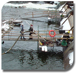 Two workers on a catwalk in a dam using industrial vacuum hose to skim oil from water